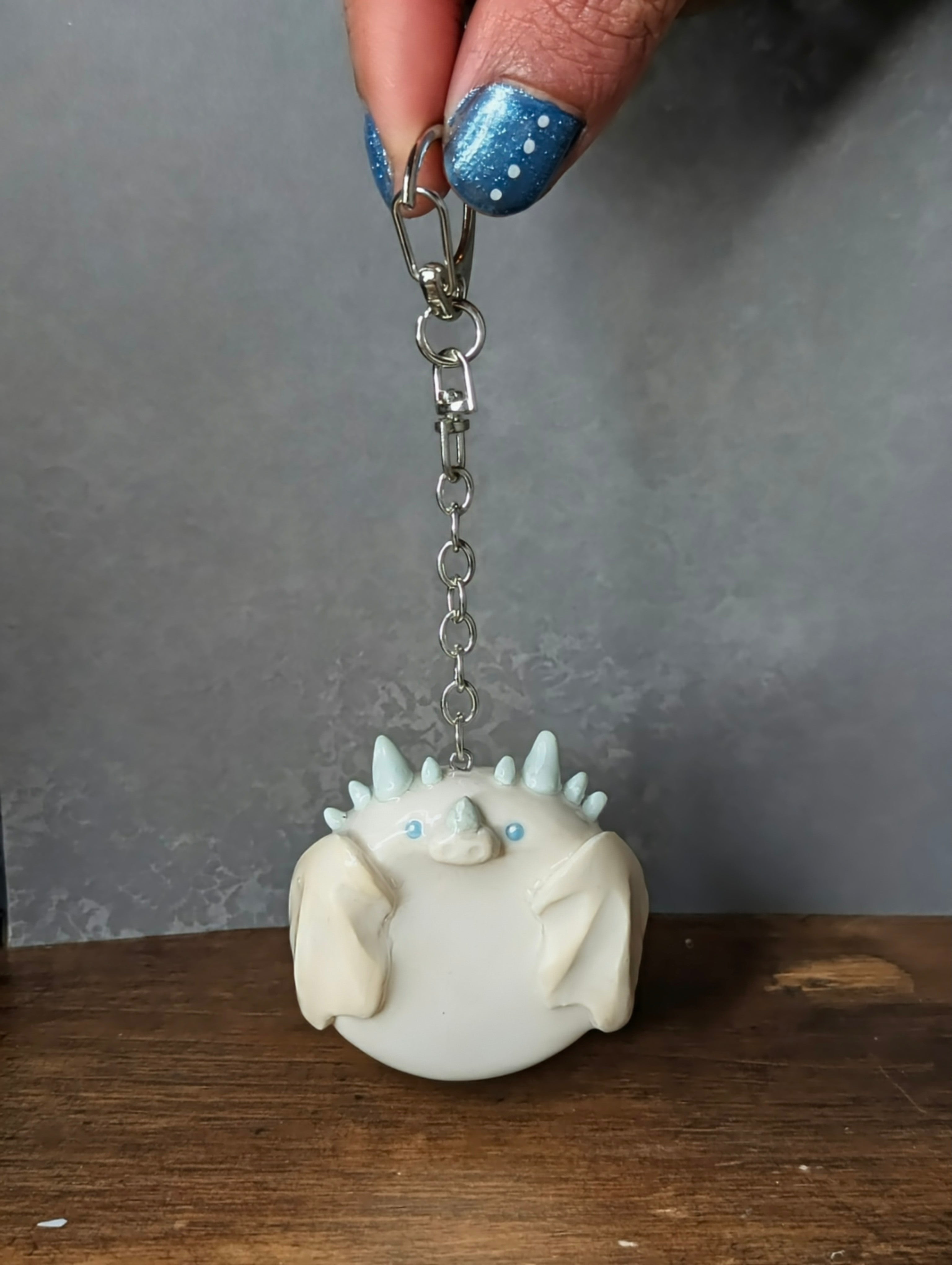 White monster-shaped keychain held by a hand with blue nail polish against a gray background Handcrafted ice dragon worry stone in pale blue crystal gemstone